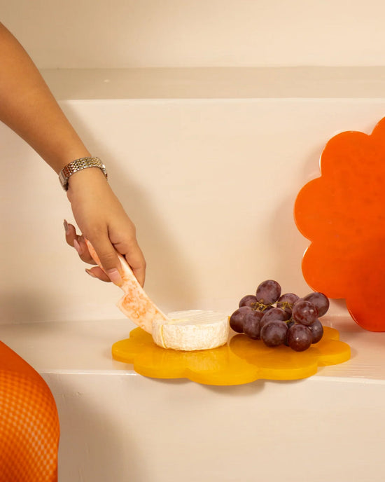 A hand with a cheese knife hovers over cheese and grapes on the Casa Amarosa 9in Scallop Resin Cheese/Charcuterie Board in Mustard, shaped like a yellow flower.