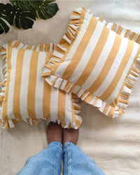 Two Casa Amarosa Golden Yellow Ruffle 16x16in pillows, featuring yellow and white stripes with ruffled edges, rest on beige fabric. A barefoot person in blue jeans stands below, while a green leaf accents the upper left corner.