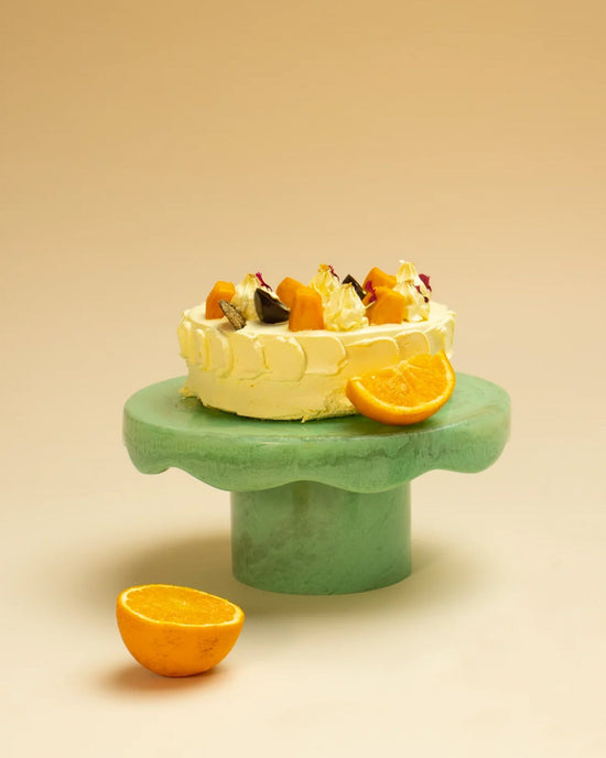 A round frosted cake with assorted fruit rests on the Casa Amarosa Scallop Resin Cake Stand in Green, with a halved orange and an orange slice beside it.