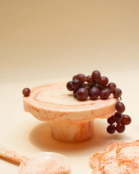 A cluster of red grapes sits on the Scallop Resin Cake Stand in Ivory Pink by Casa Amarosa, with matching marbled serving pieces nearby, all set against a neutral background.