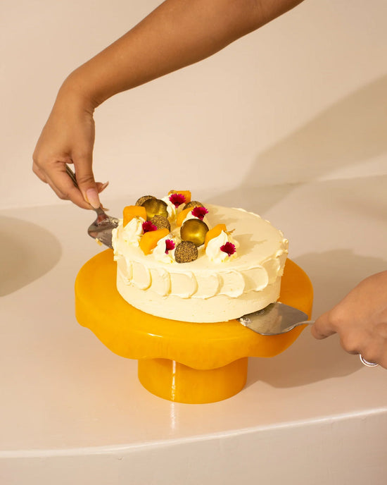 Using two cake servers, a person lifts a decorated round cake with white frosting and gold toppings from the Casa Amarosa Scallop Resin Cake Stand in Mustard.