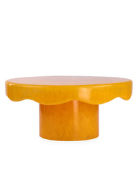 The Casa Amarosa Scallop Resin Cake Stand in Mustard features a round, yellow wavy-edged top and cylindrical base, evoking a playful design, displayed against a white background.