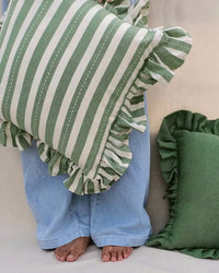 A barefoot person in loose light blue jeans holds the Casa Amarosa Striped Green Ruffle 16x16in Pillow; another green-ruffled, 100% cotton pillow from the same brand is nearby.