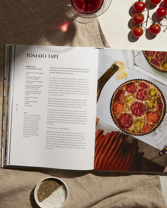 A Table by Chronicle Books is open to a French-inspired tomato tart recipe, shown with a photo of the tart. Around it are cherry tomatoes, red wine, and bowls of salt and pepper.