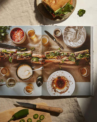 An open copy of A Table by Chronicle Books displays French recipes beside a long sandwich with garnishes; drinks, sauce, and sliced jalapeños are arranged around the book on a light tabletop.