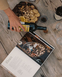 A hand holds a bottle of alcohol above the open French cookbook A Table by Chronicle Books, with a bowl of mixed nuts and snacks arranged on a wooden table.