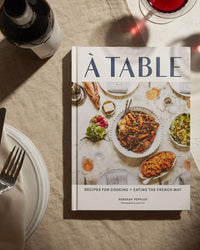 A Table by Chronicle Books, a French cooking cookbook, rests on a table beside a wine glass, fork, knife, and plate. The cover features an enticing spread of French cuisine.