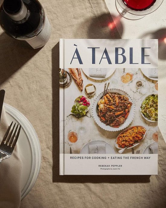 A Table by Chronicle Books, a French cooking cookbook, rests on a table beside a wine glass, fork, knife, and plate. The cover features an enticing spread of French cuisine.