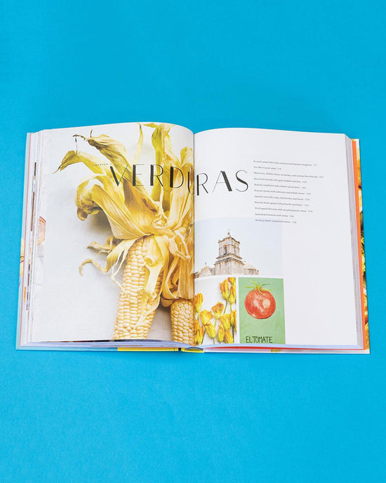 An open book, Ama by Chronicle Books, rests on a blue surface showing images of corn, a building, and a tomato with VERDURAS in Spanish—hinting at Tex-Mex recipes inspired by regional Mexican cuisine.