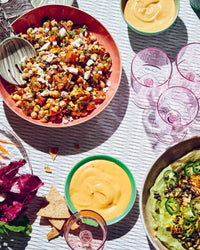 A table set with a chickpea salad from Epic Salads by Chronicle Books, green dip and vegetables, two bowls of orange dip, pink glasses, and chips scattered on a striped tablecloth.