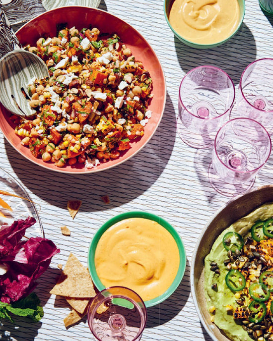 A table set with a chickpea salad from Epic Salads by Chronicle Books, green dip and vegetables, two bowls of orange dip, pink glasses, and chips scattered on a striped tablecloth.