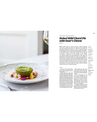 A small green tart, inspired by Italian desserts, is served on a floral plate beside a fork; Recipes from Sicily by Chronicle Books appears in the scene, while a blurred glass of white wine sits in the background.