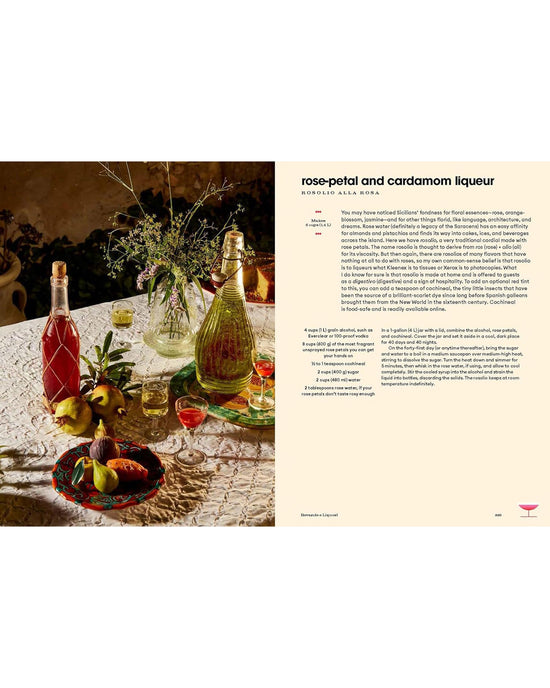 A table with a lace tablecloth holds glass bottles, colorful drinks, fruit, vases of flowers, and an open copy of Sicily, My Sweet by Chronicle Books displaying a rose-petal and cardamom liqueur recipe.