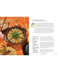 A cast iron pot with sardine fillets over rice and herbs rests on a red marble table next to chopsticks, a glass of water, and an open copy of Tin to Table by Chronicle Books.