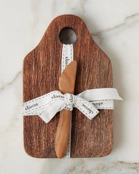 The etúHOME Dutch Small Cheese Board w/ Spreader in Natural features a handy top hole, is tied with a white ribbon, and comes with a wooden spreader—an ideal hostess gift, beautifully presented on light marble.