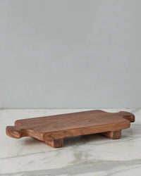 The Normandy Wood Riser Footed Tray, Medium by etúHOME features short legs and end handles, crafted from Acacia wood and elegantly displayed on a white marble surface with a gray backdrop.