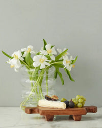 A clear glass vase with white flowers sits beside the etúHOME Normandy Wood Riser Footed Tray, Small, which holds cheese, figs, and green grapes on a white surface against a gray wall.