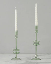 The etúHOME Sage Glass Candlesticks Set of 2 - Large features handmade, green recycled glass holders displaying white twisted candles on a marble surface with a light gray background.