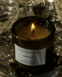 A dark green Coriandoli Candle by Flamingo Estate burns gently, its vegetable wax casting a warm glow amid glassware and a metallic surface, adding elegance to any setting.