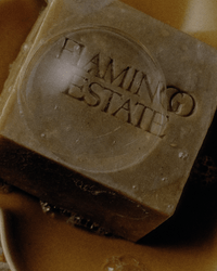 A close-up of Flamingo Estates Manuka Honey Bar Soap, a brown bar with the brand engraved, resting on a beige soap dish with visible bubbles.