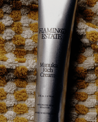 A silver tube labeled Flamingo Estate Manuka Rich Cream, a hydrating cream featuring Manuka honey, sits atop textured yellow and white woven fabric.