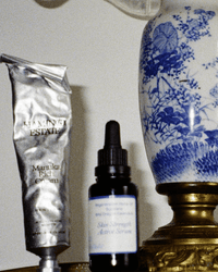A metallic tube of Flamingo Estate Manuka Rich Cream sits beside a porcelain lamp with blue floral designs and a brass base.