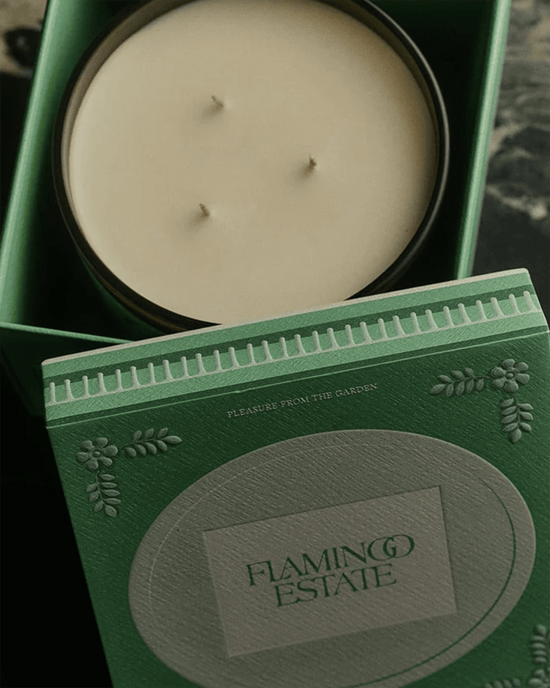 A green box holds Flamingo Estate’s The Tomato XL Candle, crafted from vegetable wax, beside its matching lid embossed with decorative patterns and the phrase Pleasure from the Garden.