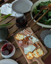 A wooden table set with a charcuterie board, crackers, cured meats, cheese, almonds, two glasses of red wine on graf lantz Bierfilzl Merino Wool Felt Round Coasters in Noir, salad bowl, and plates—perfect for sustainable home decor enthusiasts.