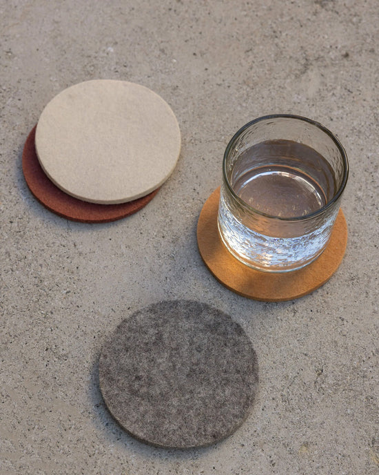 A glass of water rests on a graf lantz Bierfilzl Merino Wool Round Coaster Multi in Mesa (4 Pack), with grey and beige coasters beside it, all on concrete—a stylish, sustainable décor accent.