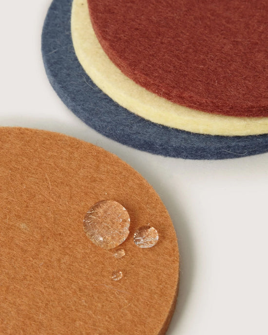 The graf lantz Bierfilzl Merino Wool Round Coaster Multi in Yucca - 4 Pack features brown, blue, yellow, and red sustainable coasters stacked, with water droplets on the brown coaster in front.