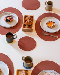 A breakfast table set with mugs of coffee on graf lantz Bierfilzl Merino Wool Round Coaster Multi in Yucca (4 Pack), plates of pastries, a tray of croissants, and red circular placemats on a white surface.