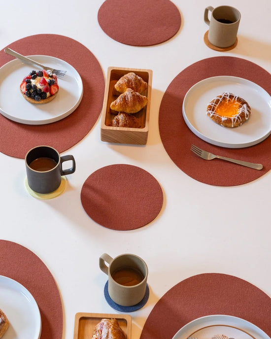 A breakfast table set with mugs of coffee on graf lantz Bierfilzl Merino Wool Round Coaster Multi in Yucca (4 Pack), plates of pastries, a tray of croissants, and red circular placemats on a white surface.