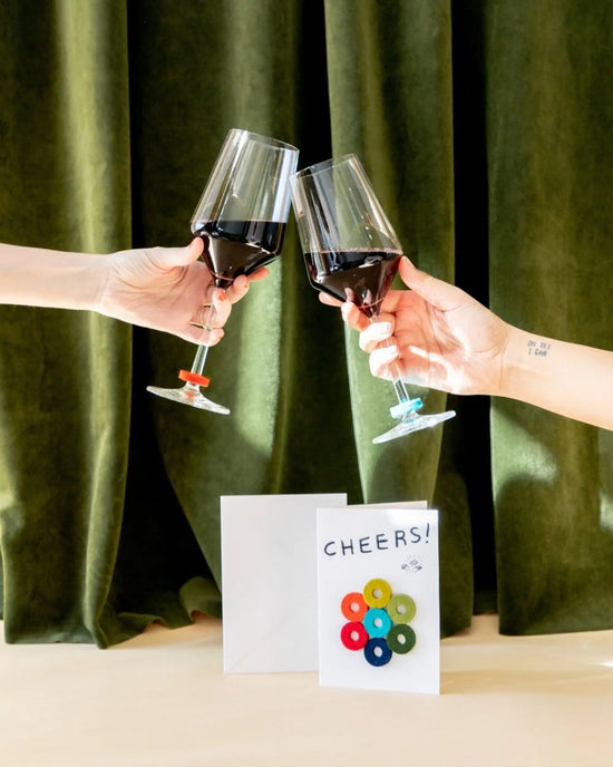 Two hands clink red wine glasses before green curtains. On the table, a graf lantz Cheers Wine-Otes Merino Wool Felt Wine Marker Note Card and playful wine charms add a festive touch to the celebration.