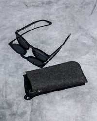 A pair of black sunglasses sits next to the graf lantz Merino Wool Felt Anzen Eyeglass Sleeve in Charcoal, combining stylish eyewear and premium protection on a sleek gray concrete surface.