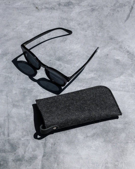 A pair of black sunglasses sits next to the graf lantz Merino Wool Felt Anzen Eyeglass Sleeve in Charcoal, combining stylish eyewear and premium protection on a sleek gray concrete surface.