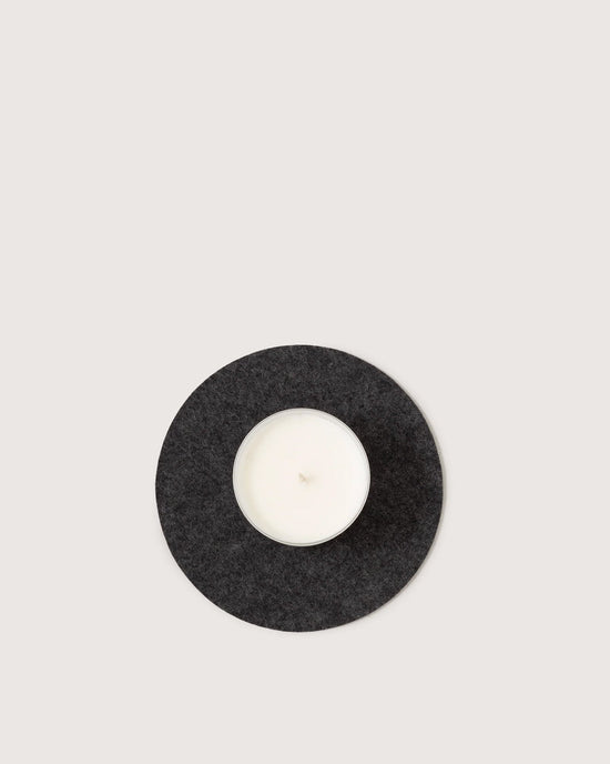 A white candle in a glass holder rests on the graf lantz Round Merino Wool Felt Trivet 8 in Charcoal—an elegant, sustainable kitchen accessory.