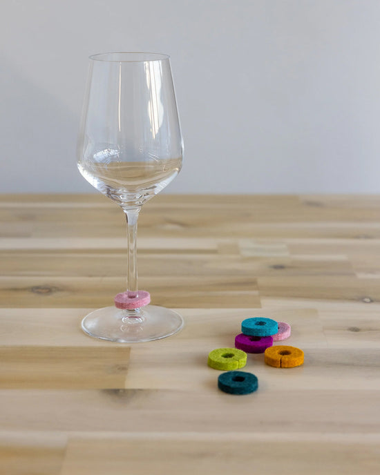 A graf lantz Salud Wine-Otes Merino Wool Felt Wine Marker Note Card rests on a wooden table beside colorful felt rings, perfect as unique wine charms or a thoughtful host gift.