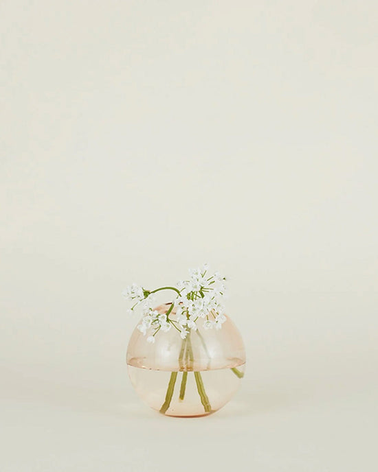 The Aurora Sphere Vase in Blush by Hawkins New York features handblown transparent glass with pale pink tones, holding delicate white flowers and green stems against a simple light background.