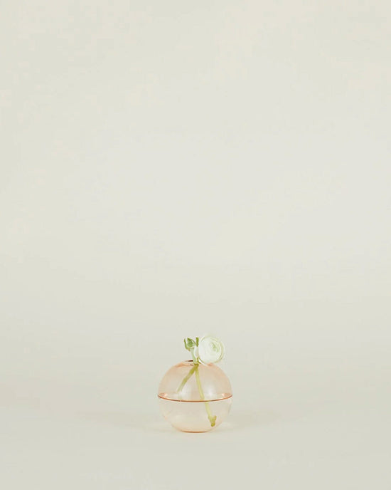 The Aurora Sphere Vase in Blush by Hawkins New York, a round translucent pink handblown glass piece with a green leaf and stem detail, is displayed centered on a plain light background.