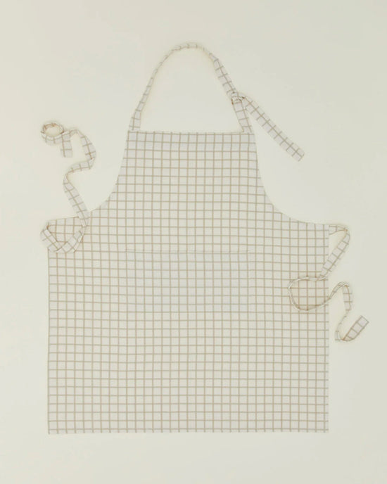 The Hawkins New York Essential Check Apron in Ivory/Flax features a white cotton design with thin brown grid lines, plus adjustable neck and waist ties for the perfect fit—a thoughtful kitchen gift for any cooking enthusiast.