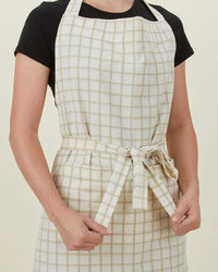 Person wearing Hawkins New York Essential Check Apron in Ivory/Flax, tied at the waist over a black t-shirt and holding the adjustable straps—a thoughtful kitchen gift.