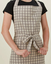 A person wearing a black t-shirt and the Hawkins New York Essential Check Apron in Light Grey/Dark Grey, tied at the waist, stands against a plain background.