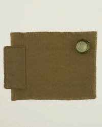 An Essential Dinner Napkin in Olive by Hawkins New York is placed on the left of a brown placemat, with a clear drinking glass at the top right, all set against a plain white background.