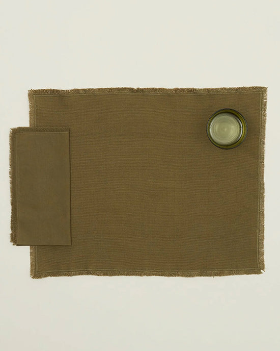 An Essential Dinner Napkin in Olive by Hawkins New York is placed on the left of a brown placemat, with a clear drinking glass at the top right, all set against a plain white background.