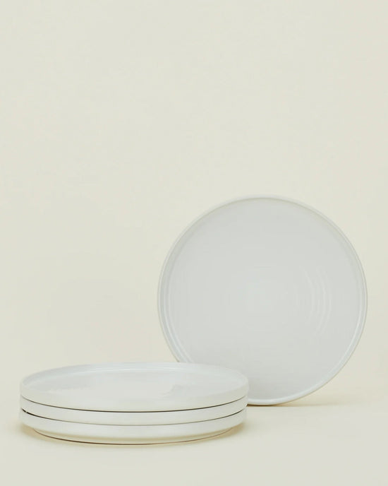 Four Hawkins New York Essential Dinner Plates in Bone are shown—three stacked and one upright behind them—displayed against a simple background; this sleek set of 4 stoneware plates is perfect for any table setting.