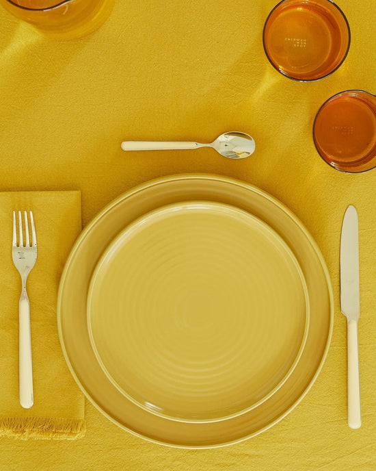 A table setting with an Essential Dinner Plate in Mustard from Hawkins New York (set of 4), on a yellow tablecloth, paired with white-handled cutlery, a yellow napkin, and two amber drinking glasses.
