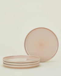 Four Essential Dinnerware in Blush plates by Hawkins New York are stacked with a matching plate standing upright behind them. This 16-piece stoneware set is both stylish and dishwasher safe, shown against a plain off-white background.
