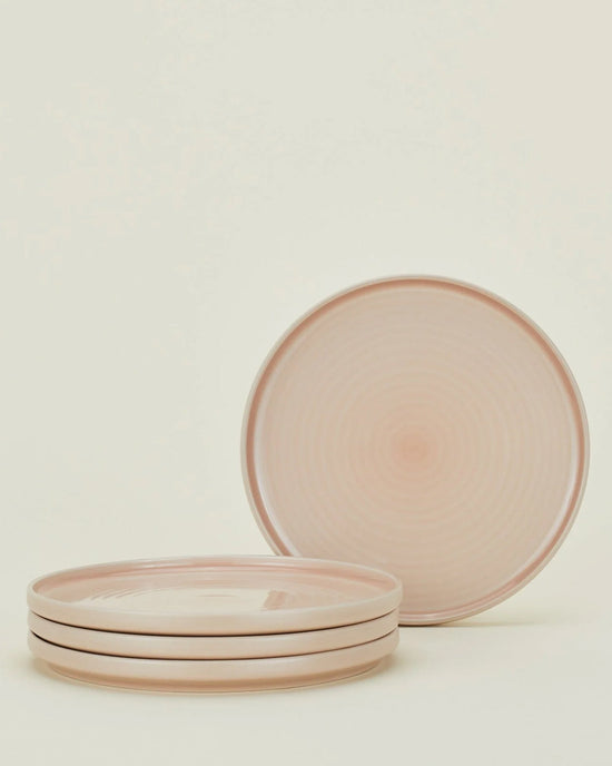 Four Essential Dinnerware in Blush plates by Hawkins New York are stacked with a matching plate standing upright behind them. This 16-piece stoneware set is both stylish and dishwasher safe, shown against a plain off-white background.