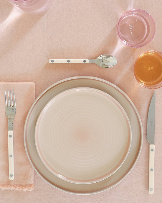 The Hawkins New York Essential Dinnerware in Blush, a 16-piece stoneware set, features plates and bowls on a pink tablecloth with flatware and two glasses. This stylish, dishwasher safe set is perfect for everyday dining.