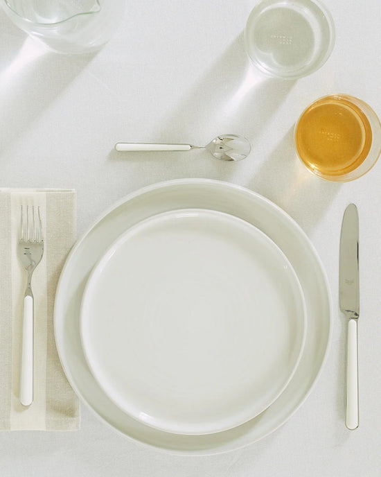 A neatly arranged place setting displays Hawkins New Yorks Essential Dinnerware in Bone—16 Piece Set: two stacked white plates, fork, knife, spoon, napkin, a water glass, and an amber drink on a crisp white tablecloth.